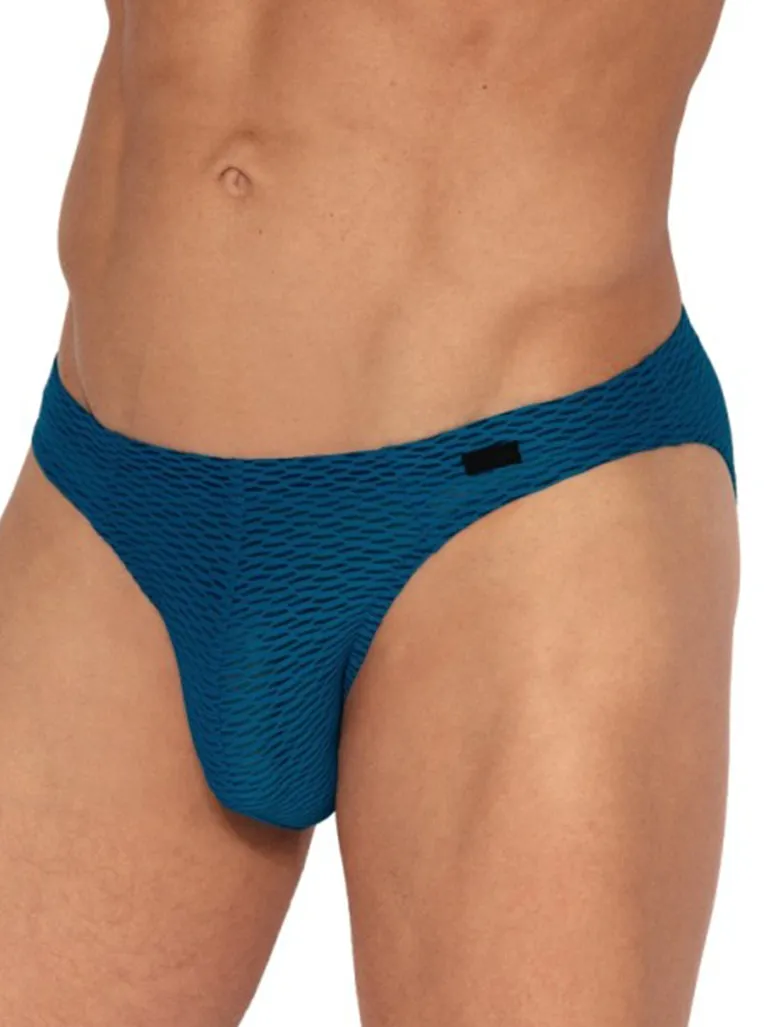 Brazilbrief Olaf Benz RED2564 Teal: ligereza visual y termorregulación El Brazilbrief Olaf Benz RED2564 Teal está pensado para quien busca una prenda de ropa interior masculina breve, ultraligera y con una imagen mucho más fresca que la de un slip liso convencional. El tono teal aporta un azul verdoso vivo y muy actual, mientras que la estructura tipo panal le da al conjunto una lectura táctil y visual mucho más trabajada que la de una superficie plana. Lo mejor de esta referencia es que el diseño no depende solo del color. El tejido con microperforaciones ayuda a que la prenda resulte excepcionalmente transpirable en el uso diario, y la construcción breve del Brazilbrief mantiene una línea limpia y compacta bajo la ropa. Si te funciona este tipo de patrón anatómico, aquí puedes ver más slips de hombre con distintos materiales, tiros y acabados. Qué aporta un Brazilbrief frente a otros slips clásicos Corte breve: reduce estratégicamente el tejido en la cadera, dejando una línea más despejada que la de un slip de mayor cobertura. Menos volumen: el patrón va al grano, evitando pliegues y ayudando a que la prenda no se marque bajo el pantalón. Perfil limpio: es una opción fundamental si buscas un interior ligero que te ofrezca total libertad de movimiento en la pierna. Tejido técnico 82/18: microfibra, transpirabilidad y frescura La mezcla de 82% poliamida (microfibra) y 18% elastano encaja a la perfección con este formato anatómico porque aporta elasticidad, una adaptación impecable y una sensación de uso que casi no se nota. En esta serie RED2564, la estructura de panal del tejido favorece una circulación del aire continua, siendo la opción definitiva para los días más cálidos o para jornadas prolongadas. Por qué el color Teal funciona tan bien en la serie RED2564 Aquí el color no se limita a decorar. El tono teal (azul verdoso) da un punto mucho más vibrante y visible que el de un slip neutro tradicional, pero sigue manteniendo una estética limpia y sofisticada. Sumado al dibujo tipo panal que genera sutiles juegos de luces y sombras, el resultado es una prenda con una fuerte identidad sin caer en estampados estridentes. Dudas frecuentes sobre el Brazilbrief RED2564 ¿Qué diferencia hay entre un Brazilbrief y un slip clásico? El Brazilbrief recorta el tejido lateral e inferior, dejando una línea anatómica más limpia, con menos volumen y una lectura visual más atrevida y breve. ¿Cómo es el color teal? Es un azul verdoso vivo y profundo, visualmente más fresco y energético que un azul marino o un tono neutro convencional. ¿Qué aporta exactamente la estructura panal? Funciona como un sistema de termorregulación pasiva. Da una lectura más ligera y trabajada al tejido, además de facilitar la evacuación del calor corporal. ¿Es buena opción para uso prolongado? Absolutamente. Precisamente uno de sus mayores puntos fuertes es combinar esa ligereza y transpirabilidad extrema para garantizar el confort durante muchas horas. Composición y Cuidados Materiales: 82% poliamida 18% elastano Mantenimiento: Lavado a máquina (máx. 40 ºC). No usar lejía bajo ningún concepto. No secar en secadora. Planchar a temperatura media si fuera estrictamente necesario. Limpieza en seco permitida (excepto con tricloroetileno). Consejo XXXMadrid: Esta referencia tiene mucho sentido si buscas un slip de hombre breve, excepcionalmente fresco y fácil de llevar en el día a día, pero con un trabajo superior en la textura (panal) y el color. Si dudas entre dos tallas o prefieres un ajuste menos compresivo en la cintura, te recomendamos elegir la superior.