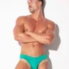 Modelo con brazos cruzados apoyado en la pared, bañador mini slip Code 22 Amsterdam verde